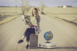 The girl is sitting on a suitcase on the road and looking at the map deciding how to handle delays when relocating, and ext to her is another suitcase with a world globe on it, and in the distance behind her are houses, trees, and grass meadows.