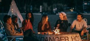 Young people sit gathering around a fire pit on the beach while night falls, laughing, talking and having fun.